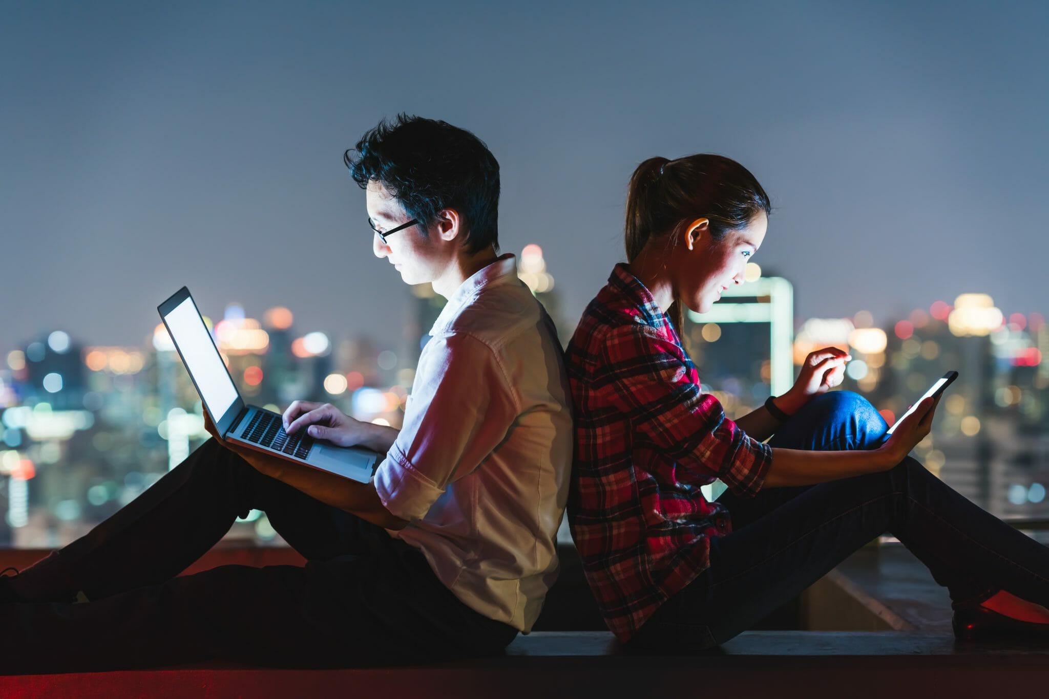 Two people sitting back to back staring at electronic devices